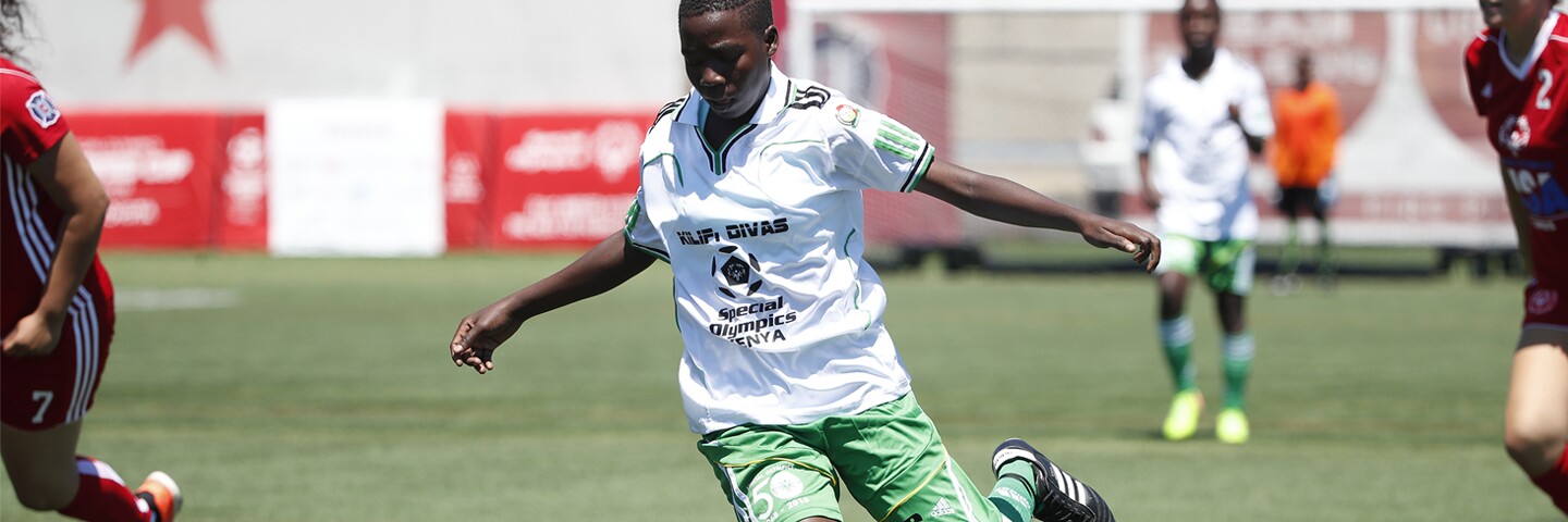 Female athlete on the pitch kicking the ball while the opposing team surrounds her. 