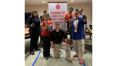 Group of seven athletes standing in front of a sign that reads, Changing the world is a contact sport, game on. Some of the athletes are holding Bocce balls. 