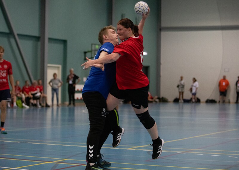 A woman wearing a red jersey jumping, holding a ball  colliding with a man on the opposing team, wearing a blue jersey