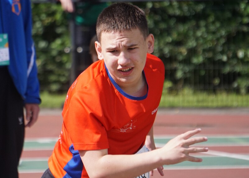 A young man in a red sports jersey standing on a sports track holds his hands in front of him in a gesture suggesting he has just thrown something forward. 