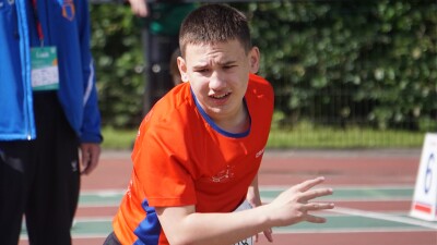 A young man in a red sports jersey standing on a sports track holds his hands in front of him in a gesture suggesting he has just thrown something forward. 