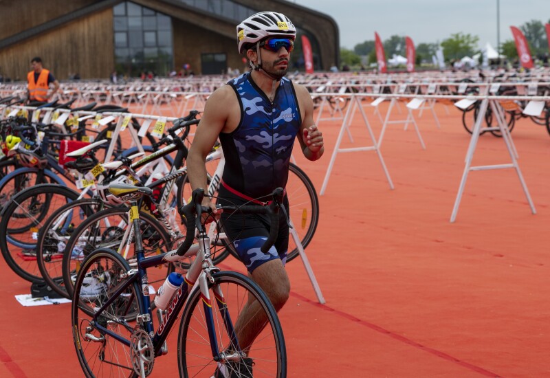 an athlete in a blue camouflage uniform and a bike helmet walks alongside his bicycle