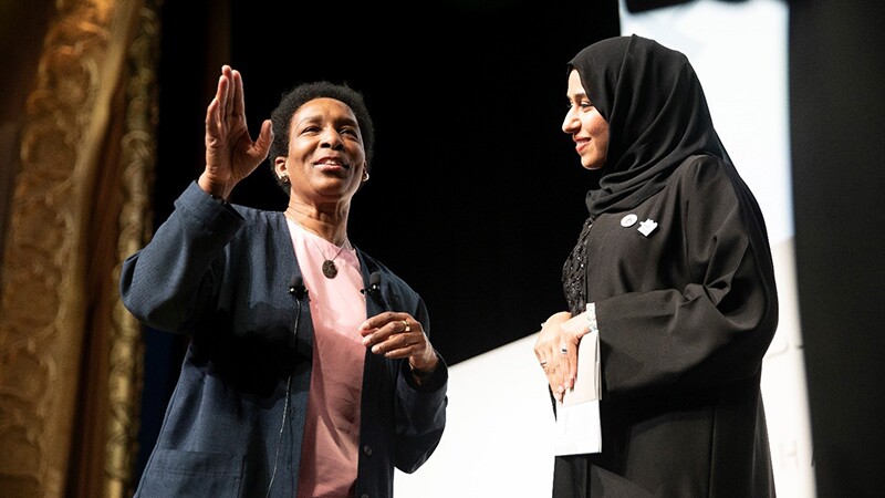 Two women with microphones standing on stage; the woman on the left (Loretta) is speaking; woman on the right is attentive. 