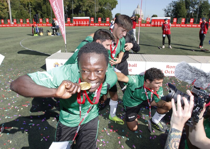 Young man on a football pitch, in front of a podium celebrating with four teammates and biting a golden medal