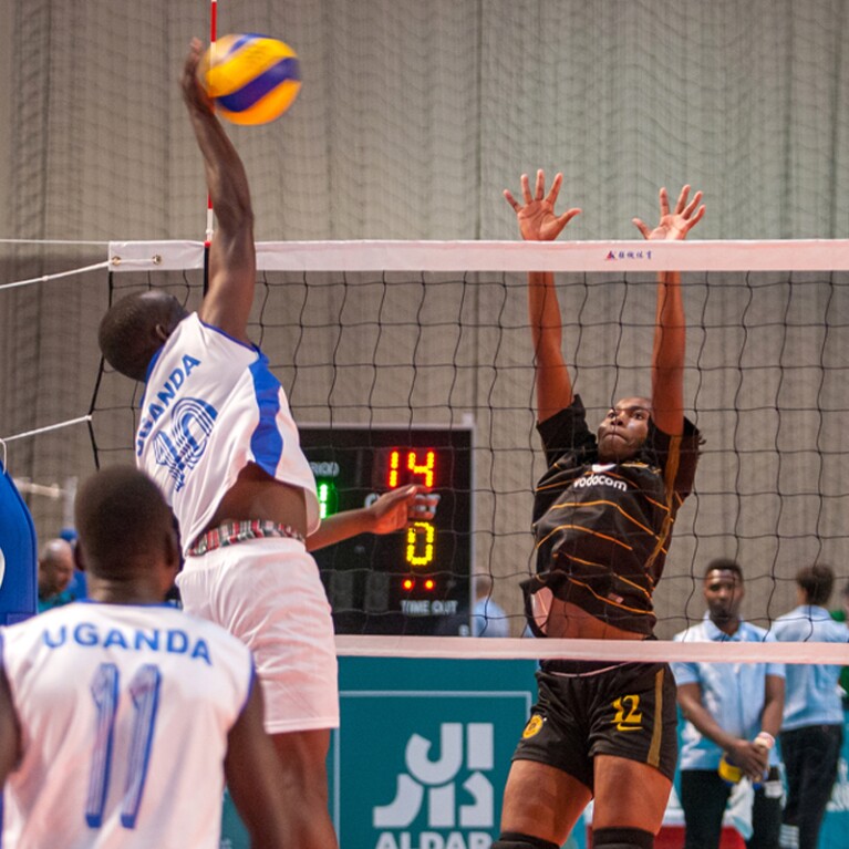 In a volleyball game, a young man is jumping to spike the ball and a young woman jumps to block it. 