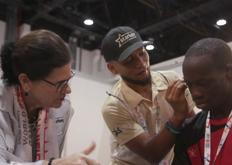 Athlete is receiving an ear exam and having his hearing aids put on by a volunteer as a medical professional observes.