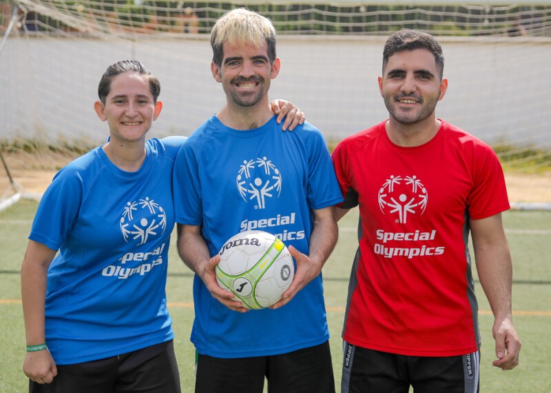 Two men, one wearing a red shirt and another a blue one, and a woman wearing a blue shirt pose for the camera while the man in the middle holds a football