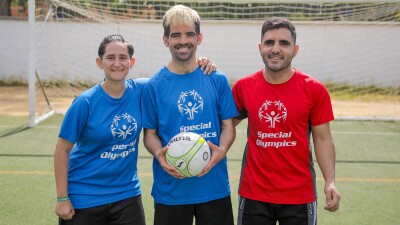 Two men, one wearing a red shirt and another a blue one, and a woman wearing a blue shirt pose for the camera while the man in the middle holds a football