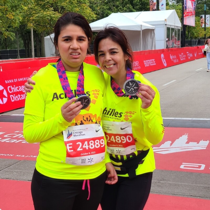 Two women stand at the finish line of a marathon, showing off their medals. 