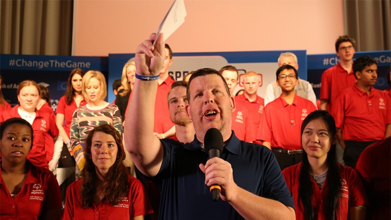 Man speaking into a microphone he's holding in his left hand and his right hand up holding note cards. Spectators are behind him. 