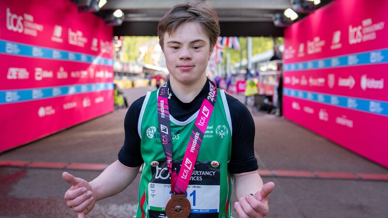 A young man wearing a green sports bib and with a medal around his neck faces the camera holding his thumbs up. 