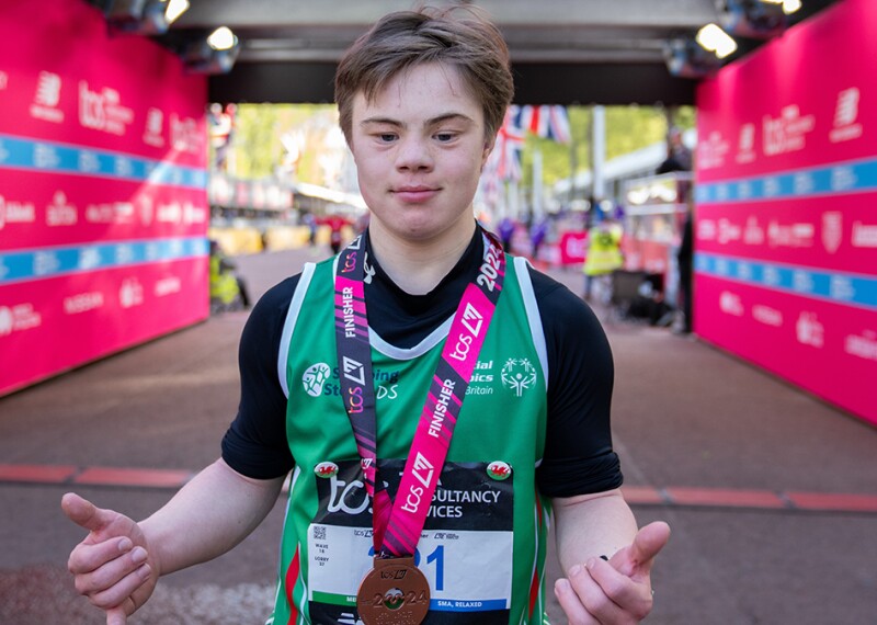 A young man wearing a green sports bib and with a medal around his neck faces the camera holding his thumbs up. 