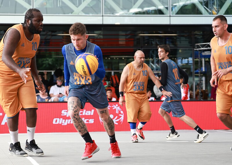 Five basketball players on court, three in orange kits and two in grey with fans and signage in the background. 