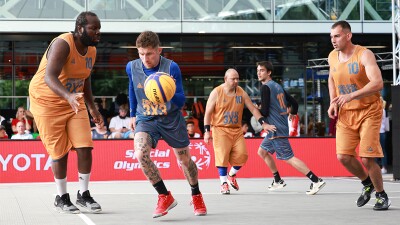 Five basketball players on court, three in orange kits and two in grey with fans and signage in the background. 