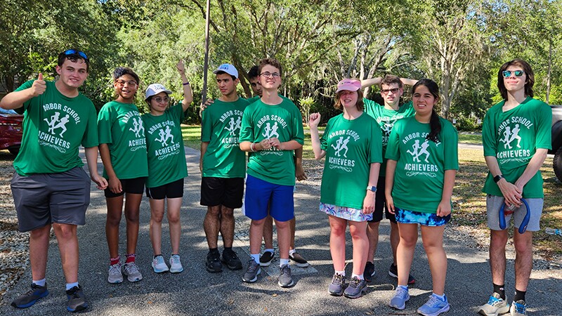 The photo shows 10 individuals wearing matching green shirts that say "Arbor School Achievers". They are standing outside and smiling for a photo. 