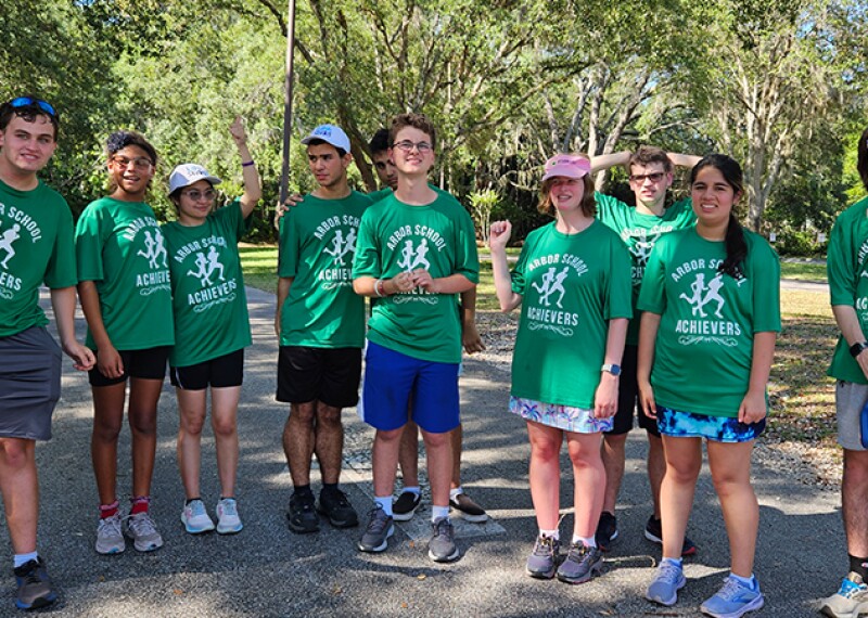 The photo shows 10 individuals wearing matching green shirts that say "Arbor School Achievers". They are standing outside and smiling for a photo. 