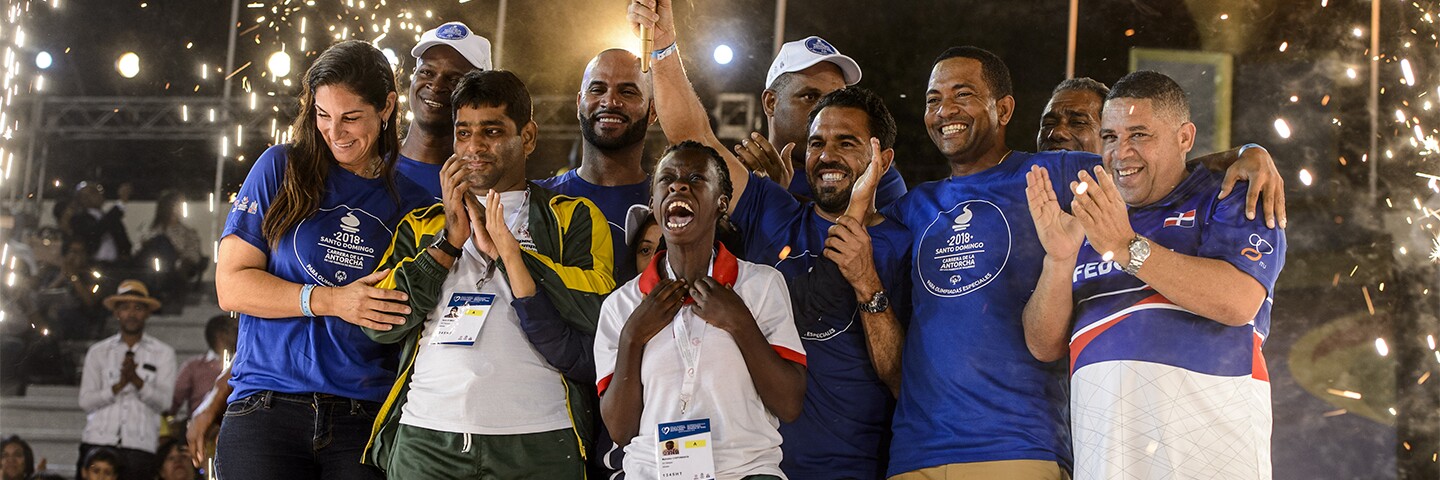 A group of athletes and representative celebrate as fireworks go off in the background. 