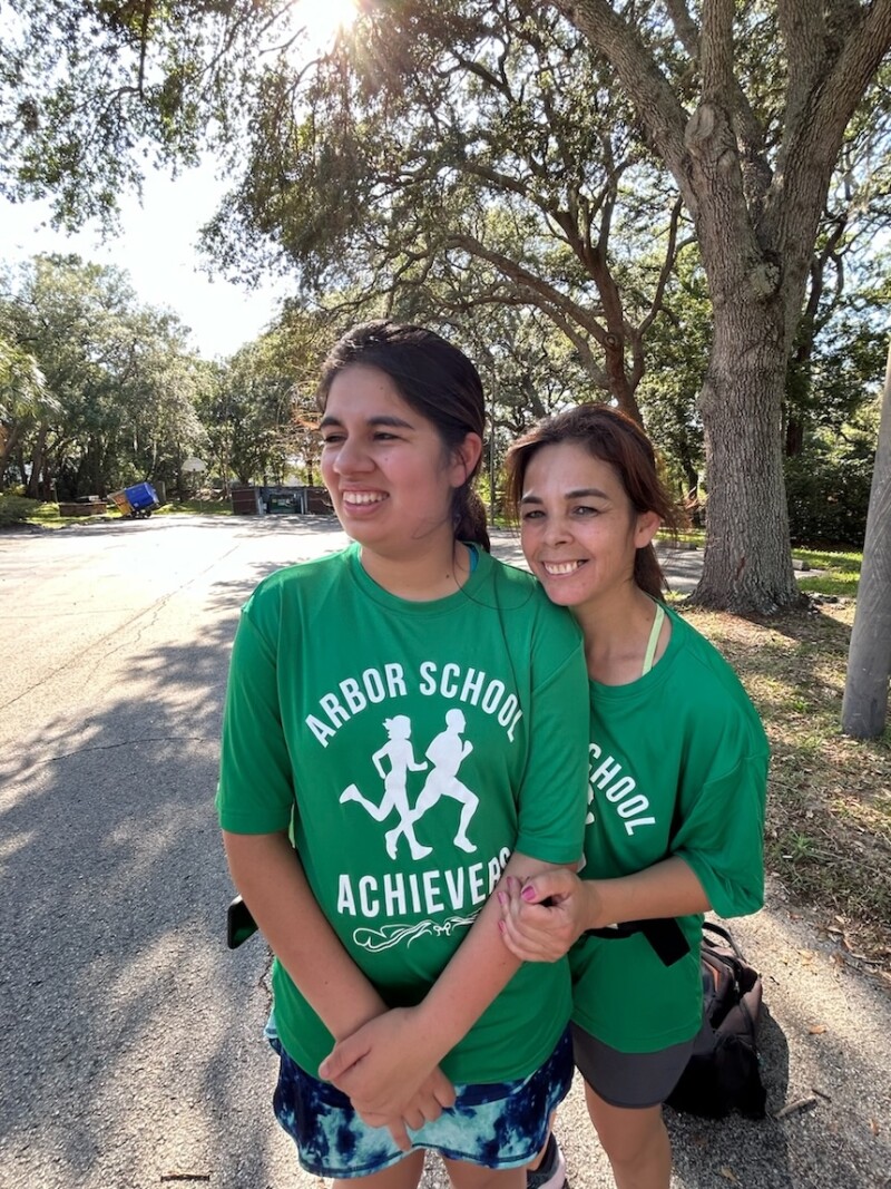 Two women wearing matching green shirts that say "Arbor School Achievers" are posing for a photo outside. 