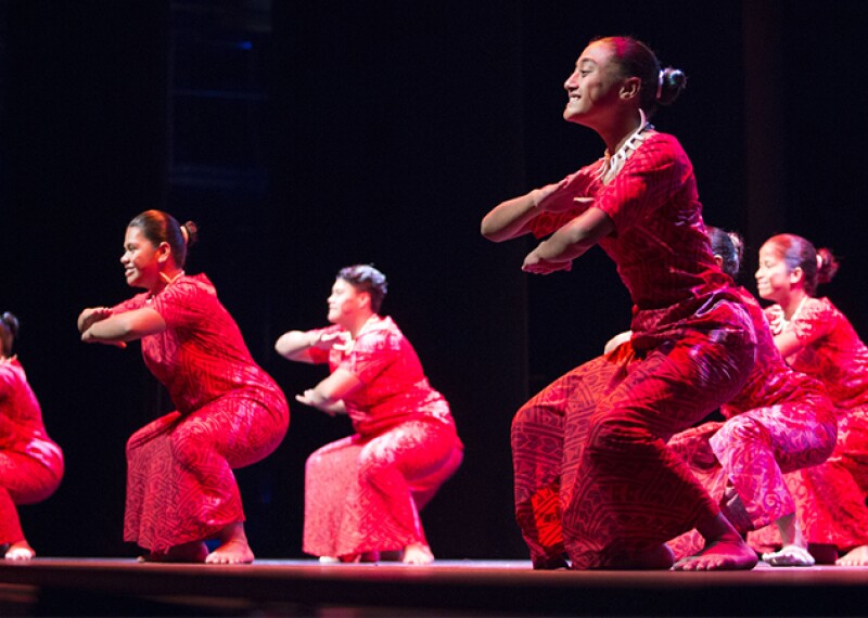 Six dancers on stage wearing red shirts and sarongs.