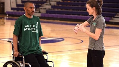 Man in wheelchair holding himself up and woman is timing him with a stopwatch. 