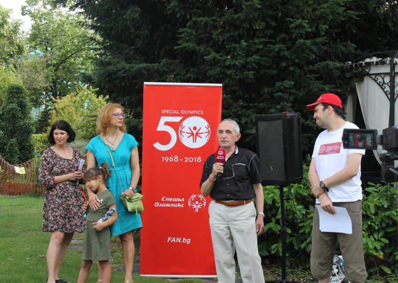 Two women, one girl and two men stand in front of a Special Olympics banner in a garden setting. The man is holding a red microphone.