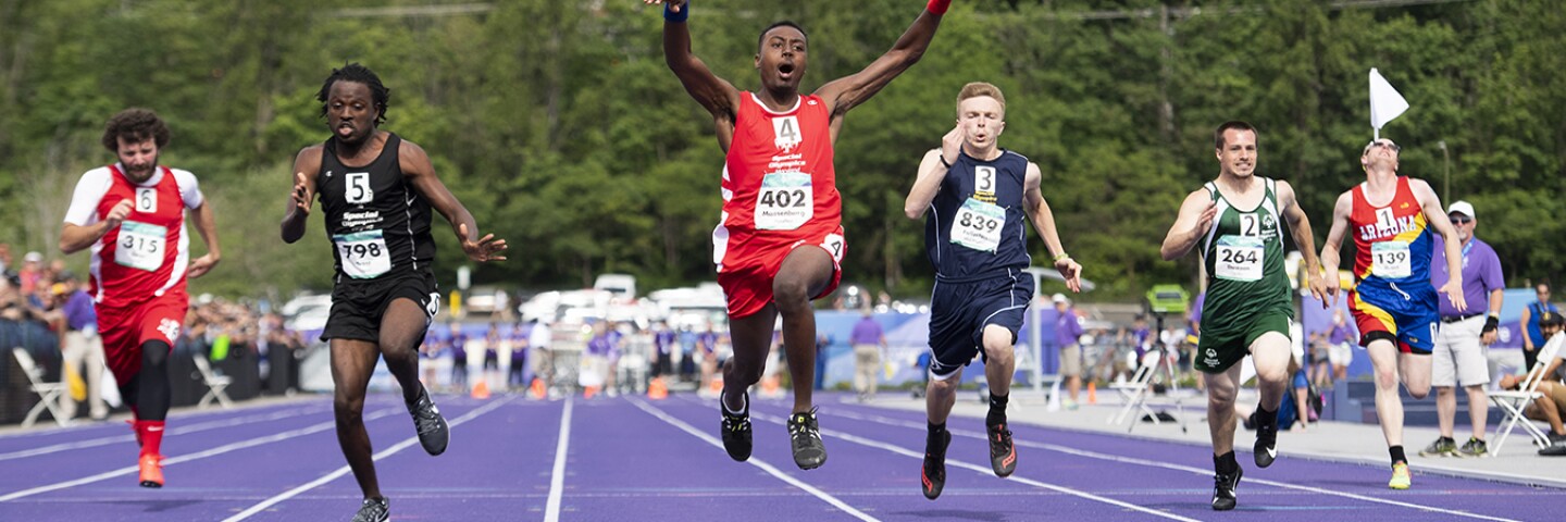 A group of six male runners running on a track with spectators in the background. 