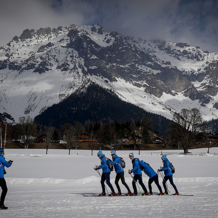 5 Cross Country Skiers skiing across the snow as a coach watches. 