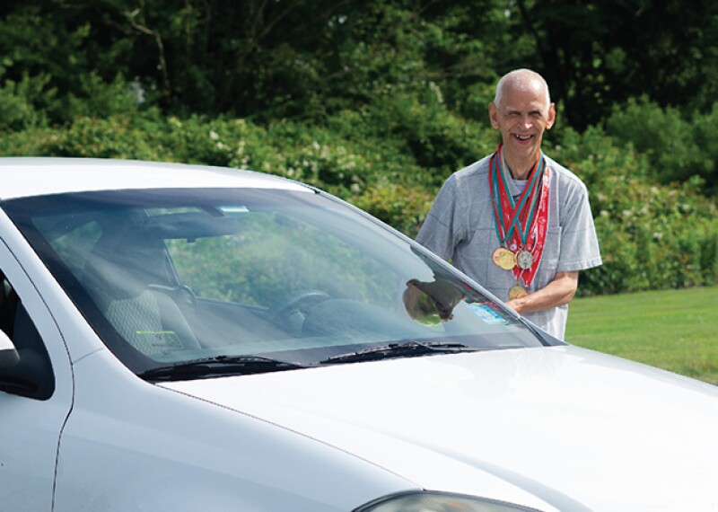 Athlete with medals around his neck standing beside a white car. 