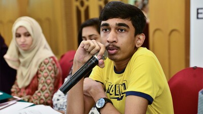 Young adult male in a yellow shirt sitting at a table talking into a microphone. Two females can be seen in the background. 