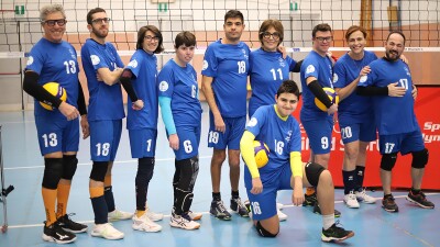 A group of volleyball players stand together as a team wearing blue kits in front of a volleyball net. 