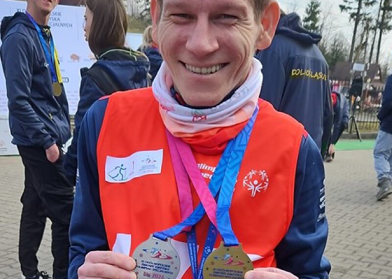 Konrad is wearing a red Special Olympics jersey and smiles at the camera while holding his medals.